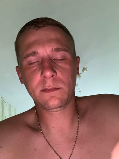 A low-angle, close-up, indoor, medium-shot, of a shirtless man with a gold chain around his neck. He has short brown hair and his eyes are closed. The background is a plain wall with some light fixtures. The lighting is warm and reddish. It is impossible to determine the exact location within the city of Minsk, Belarus, based solely on the provided image.