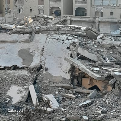 Debris and rubble are piled high, revealing remnants of a destroyed building. Concrete slabs, shattered bricks, and loose gravel dominate the scene. In the background, parts of an adjacent, seemingly undamaged building are visible, showcasing windows and balconies. The destruction contrasts starkly with the standing structure, indicating a catastrophic event. A "Galaxy A14" watermark appears in the corner.