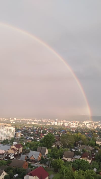 This media file captures a panoramic view of a city under a sky adorned with a vibrant rainbow. The scene is a blend of urban development and lush greenery. In the foreground, a dense residential area with numerous houses, primarily single-family homes with pitched roofs, is visible. These houses are nestled amongst abundant trees, creating a suburban feel. Some homes have visible garages or carports.
Further in the distance, the urban landscape transitions to a more densely built-up area with apartment buildings and taller structures, suggesting a city center. A red and white radio tower stands out amongst the houses, indicating communication infrastructure. The overall impression of the city is one of a sprawling settlement, with a mix of older and newer buildings.
The most striking element is the prominent rainbow arcing across the sky, with its colors subtly visible against the predominantly overcast or hazy sky. The rainbow suggests a recent or ongoing rain shower, characteristic of the weather conditions. The lighting, with the warm glow diffused by the clouds and the subtle illumination of the city, indicates it is likely late afternoon or early evening.
The location is identified as Krasnoyarsk, Russia, which provides context for the architectural style of the buildings and the geographical setting, possibly with mountains or hills in the far distance, partially obscured by the haze. There are no visible people or specific events taking place; the image focuses on the landscape and the atmospheric phenomenon of the rainbow. The emotions evoked are likely awe and serenity, inspired by the natural beauty of the rainbow over the cityscape. No visible text is present in the image.