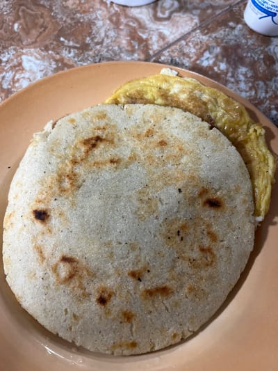 A close-up, overhead shot shows a plate of food on a tiled surface. The plate is a light orange color. On the plate are two food items: a large, circular white corn patty, slightly browned in spots, and a folded omelet, light yellow with some darker brown edges. The tiled surface beneath the plate has a marbled pattern of brown, pink, and white. A white bottle with blue lettering is partially visible in the upper right corner. The image appears to be taken in Juan Griego, Venezuela.