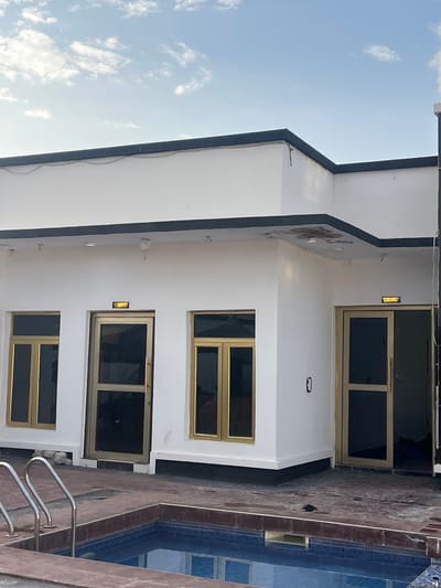 The image depicts the exterior of a modern building in Jauro Yinu, Nigeria, with a small swimming pool in the foreground. The building is painted white with black trim along the roofline and base. It features three large windows with gold frames and dark tinted glass, and two doors, also with gold frames and glass panels. One door is a single panel, while the other appears to be a double door or has a large glass panel on the side. Above one of the doors, a rectangular sign with a yellow light is visible. A security camera is mounted on the exterior wall. The swimming pool is tiled with blue mosaic tiles and has a dark red coping. Stainless steel handrails are installed on the side of the pool. The surrounding ground is paved with dark tiles. The sky above is blue with scattered white clouds. There are no people visible in the image.