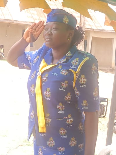A medium, full-body shot depicts a woman in uniform offering a salute. She has dark hair pulled back and is wearing a blue uniform imprinted with "GBN" and a symbol. A yellow sash is draped around her neck. She also wears a blue cap with a similar insignia. Her right hand is raised to her forehead in a salute.
The setting appears to be outdoors, with buildings and trees visible in the background. The location is likely in Akwanga, Nigeria. Other features are a generator set.