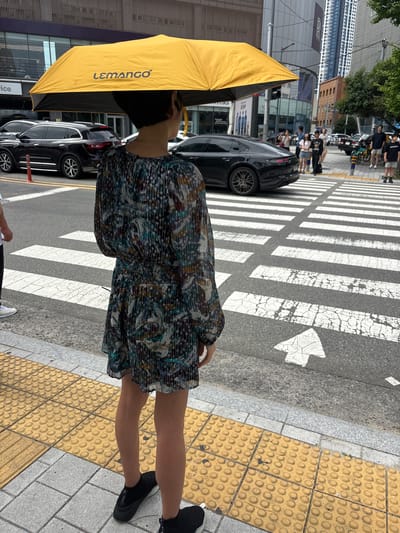 The image shows a street scene in Busan, South Korea. A person stands on a sidewalk, holding a yellow umbrella over their head. The person is wearing a patterned dress and black sneakers. Across the street, there is a pedestrian crossing with white stripes on the asphalt. Several cars are visible on the street, including a black sedan. In the background, there are buildings of varying heights, including a tall skyscraper. People can be seen walking on the sidewalk across the street and near the pedestrian crossing. The overall impression is a typical urban environment in a South Korean city.