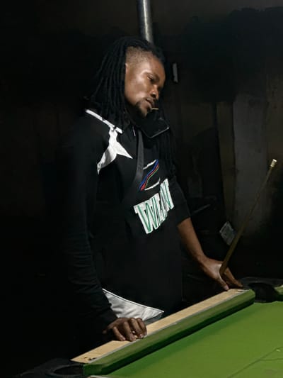 A man is playing pool in a dimly lit room. He is Black with dreadlocks and is wearing a black long-sleeved shirt with white and green lettering. He is holding a pool cue with his right hand and is guiding the edge of the pool table with his left hand. The pool table is green. There is a piece of paper or a bill stuck to the wall behind him. The location is Ogwashi-Uku, Nigeria.