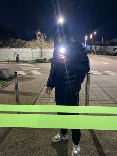 A nighttime image depicts the reflection of a lone individual standing in Hœnheim, France. The subject, visible from approximately the waist up, wears a black puffer jacket, dark pants (possibly with white stripes on the side), and light-colored athletic shoes featuring reflective elements. They are holding a smartphone, its activated flash creating a bright glare that obscures the face and upper torso. A prominent horizontal green band crosses the lower portion of the reflective surface in the immediate foreground.
Behind the reflection, two silver bollards are positioned. The background reveals a dark night sky over an urban or semi-urban street scene. To the left, a dark-colored bicycle is parked next to a curb, behind which a light-colored slatted fence runs horizontally. Beyond the fence, bushes and a small, bare-branched tree are visible. A pedestrian crosswalk is marked on the asphalt road, which stretches towards the right side of the frame. Multiple streetlights illuminate the area, with one overhead light source creating a distinct vertical lens flare above the individual's reflection. Further in the distance on the right, illuminated red and yellow signs, possibly from a commercial establishment, are visible along with a white utility van. Large, natural rocks are integrated into the landscape bordering the curb.