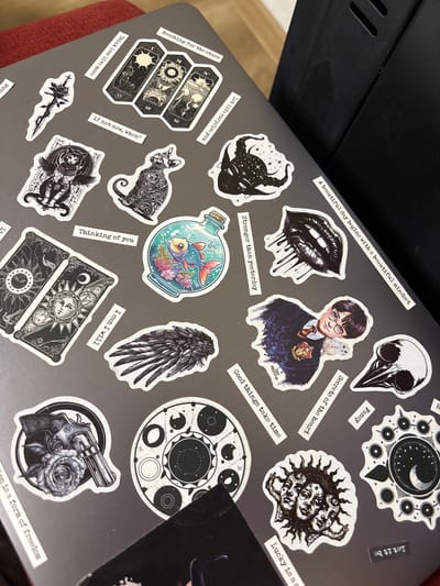 The image displays a close-up, high-angle view of a laptop lid adorned with numerous stickers, creating a personalized and eclectic surface. The laptop's lid is a dark grey color, largely obscured by the arrangement of the decals.
The stickers showcase a diverse range of artistic styles and themes, predominantly featuring monochrome or limited color palettes. Notable designs include a portrait of Harry Potter wearing his Hogwarts uniform with Hedwig perched on his shoulder, a black bird skull, a pair of stylized black lips with dripping liquid, and a horned creature's head with starry patterns. Other prominent stickers depict a colorful fish in a glass bottle, a detailed black cat, a single black feather wing, and various celestial or mystical motifs such as moons, stars, and suns, often incorporated into circular or rectangular patterns resembling tarot cards. A sticker of a revolver with a rose and a dagger with a rose are also visible.
Interspersed among the illustrative stickers are smaller, white rectangular stickers containing motivational or introspective text. These phrases include "Reaching for the stars," "Let life surprise you," "Think less live more," "If not now, when?", "A beautiful day begins with a beautiful mindset," "Stronger than yesterday," "Fancy," "Secrets of the heart," "Good things take time," "Thinking of you," "I can I will," and "Let it be." Partially visible texts read "Lucky is a s" and "on is a form of freedom." An artist's signature, "KIRYANOVA," is also visible on the Harry Potter sticker.
The laptop appears to be placed indoors, with a dark, possibly wooden, object behind it and a reddish, textured fabric (likely upholstery) visible on the lower left side, suggesting a comfortable, personal setting in Kuklen, Bulgaria. No specific activity is occurring, but the arrangement reflects the owner's personal interests and aesthetic.