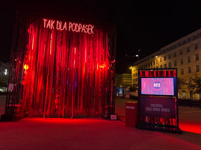 This image captures an outdoor nighttime scene in the city of Poznan, Poland, likely during the Malta Festival. In the foreground, a large, illuminated installation dominates the left side of the frame. This structure is draped with numerous red fabric strips and beaded garlands, creating a textured, curtain-like effect. Above the drapery, a neon sign in bright red letters reads "TAK DLA PODPASEK."

To the right of the main installation, a separate display features a digital screen showing the number "8212" inside a circular graphic. Below this screen is a dark sign with white text that reads "NACIŚNIJ, JEŚLI MASZ OKRES," which translates to "Press if you have a period." Adjacent to this display, a red, rectangular podium stands with the words "MAM OKRES" printed on its side.

The ground is paved with textured reddish-brown material, reflecting the red light from the installations. In the background, a multi-story building with numerous windows is visible, illuminated by streetlights. Further in the distance, elements of urban infrastructure, including cranes and other structures, are faintly discernible against the dark night sky. The overall atmosphere is one of public art and awareness, likely related to menstrual health.