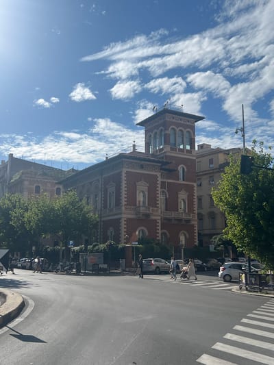 This image depicts a sunny urban street scene at the intersection of Via Saverio Mercadante and Via Po in Rome, Italy. The prominent building dominating the right side of the frame is a multi-story structure with red brick facades and light-colored classical architectural detailing, including arched windows, ornate cornices, and quoins. A distinctive, lighter-colored tower with an open loggia featuring arched windows tops the building at the corner, with satellite dishes and various antennae visible on its roof.
To the left and behind this building, other multi-story residential buildings, mostly light-colored, are visible. Some greenery and small trees grow on the roofs or upper levels of these structures.
The street itself is asphalt, curving from the foreground towards the left and center. A white zebra crossing is visible in the foreground on the right side. Strong sunlight, originating from the upper left, casts distinct shadows on the road, from trees, and from the buildings.
Several individuals are present on the street. On the right, three people are crossing the zebra crossing: a woman in a light dress pushing a baby stroller, a man in a light shirt and dark trousers, and another person partially obscured. Further to the left, a man in a dark shirt and light trousers walks towards the left side of the frame in the street. Another man is walking away from the camera on the far left pavement.
Multiple vehicles are parked along the street on both sides, including cars of various models and colors, and several mopeds or scooters on the sidewalk. Green deciduous trees with full foliage line portions of the street and obscure the lower levels of some buildings. Street furniture includes a black traffic light pole on the right, other utility poles, a blue 'P' parking sign, and a small advertising kiosk on the right pavement. The sky is bright blue with scattered white cumulus and cirrus clouds.
