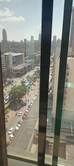 A high-angle, outdoor photographic scene captures a segment of Nairobi, Kenya, depicting a busy street and portions of the city's skyline under clear daylight conditions with scattered white clouds. The view is partially framed by two dark vertical window mullions.

In the immediate foreground, a long, sloped grey roof of a building is visible. Adjacent to this roof, a line of parked vehicles, predominantly white and red cars, occupies a street-level area. The midground is dominated by a multi-lane road with active vehicular traffic, including numerous cars of various colors and several white public transport buses moving in both directions. Pedestrians are discernible on both sidewalks bordering the road. Buildings flank the thoroughfare; a multi-story structure with a prominent grey roof and lighter facade is situated on the left, while a lighter-colored, multi-story building featuring balconies extends vertically on the right.

The distant background showcases the distinct Nairobi skyline. Notable landmarks include the Parliament Tower (KICC) with its recognizable conical roof on the far left, and the taller Britam Tower further to the right, amidst other high-rise structures and interspersed green tree cover.