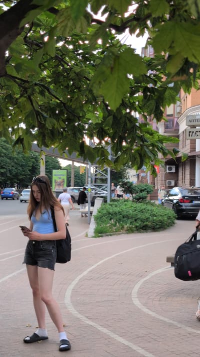 The image shows a young woman walking on a sidewalk in Poltava, Ukraine. She is wearing a light blue tank top and denim shorts, with white socks and black and grey sandals. She is carrying a black backpack and holding a smartphone in her right hand. Her left hand is resting on the backpack strap. She has long brown hair and is wearing sunglasses on her head.
The sidewalk is paved with reddish-brown bricks and has curved white lines. In the background, there are buildings, trees, and other people. One building has "ГАСТ" written on it. There is a sign that appears to be advertising a pet store, with the word "ЗООМАГАЗИН" visible. A black car is parked on the street. The weather appears to be overcast, and the time of day is likely daytime. The overall scene depicts a casual, everyday moment in an urban environment.