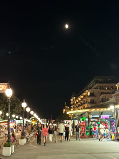 The image captures a nighttime scene along a promenade, likely located in Varna, Bulgaria. In the upper portion of the frame, a bright full moon is visible in the dark sky. The promenade is illuminated by streetlights and the glowing lights of various attractions and buildings lining the walkway.

On the left side, a neon sign spelling "SHOP" with illuminated letters is partially visible, suggesting a commercial area. Several lampposts with globes are strategically placed along the promenade, casting pools of light. The path itself is paved and appears to be a pedestrian walkway.

A variety of people are present, walking in different directions. Some are in groups, while others are individuals. They are dressed in casual attire, indicating a leisurely evening. Among the crowd, there are individuals of various ages, including children.

In the mid-ground and background, there are structures that appear to be buildings or entertainment venues. These structures are adorned with numerous lights, creating a festive atmosphere. Some of these venues seem to be arcade-style establishments, with colorful game machines visible. Further in the background, a Ferris wheel or similar amusement ride is illuminated, adding to the vibrant night scene. The overall impression is one of a lively tourist or entertainment district during the evening hours.