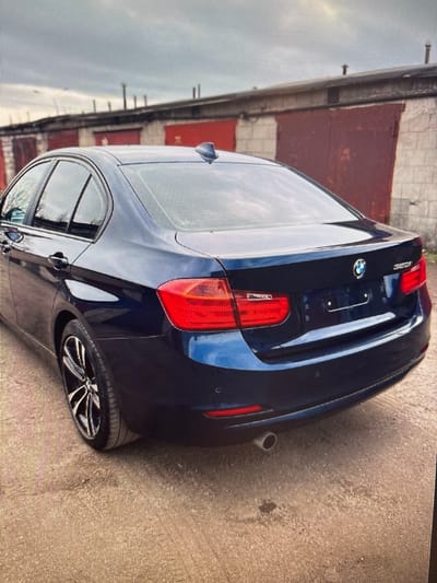 The image shows the rear three-quarter view of a dark blue BMW 320i sedan parked on a dirt and gravel surface in front of a row of garages. The garages have red metal doors and concrete walls, with some evidence of weathering and graffiti. The sky is overcast. The car is clean and appears to be in good condition, with its taillights illuminated. The license plate area is visible but the plate itself is not legible. The scene suggests an outdoor parking area, possibly in a less developed or industrial part of a city. Given the style of the garages and the general urban environment, this scene could be located in Moscow, Russia, likely in a garage cooperative or a similar storage facility.