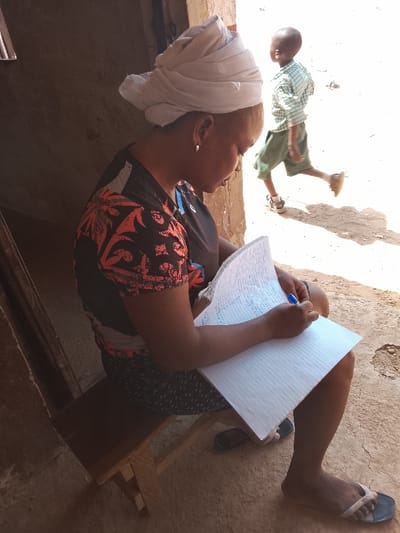 This image, captured in Akwanga, Nigeria, depicts a woman seated on a wooden bench and a child walking in the background, near a building entrance. The woman, positioned in the foreground and viewed in right profile, wears a light-colored headwrap, a dark short-sleeved top with red and orange patterns, a dark patterned skirt, and dark flip-flops. She has a silver-colored hoop earring on her visible ear. She is actively writing in an open, lined notebook with a blue pen held in her right hand, her gaze directed downwards. In the sunlit background, a child walks from right to left, wearing a light-colored, possibly checkered, short-sleeved shirt, dark green shorts, and sandals. The child is partially blurred, suggesting motion. The setting features a light brown building wall behind the woman, possibly concrete or mud, and a bright, uneven dirt or cement ground surface. Strong shadows are visible on the ground, indicating direct overhead sunlight.