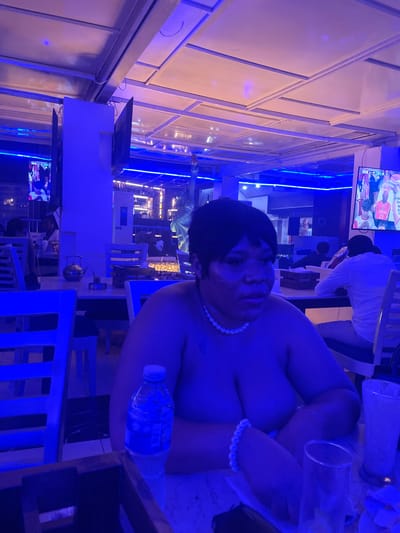 A woman with dark, short hair and a pearl necklace is seated at a table in a dimly lit establishment, bathed in blue light. She is wearing a pearl bracelet. In the foreground, to her right, are two glasses and a bottle of water. Behind her, other patrons are visible, including a man in a white shirt. A television screen displaying sports is mounted on the wall in the background. The environment appears to be a restaurant or bar with a ceiling that has a grid-like structure. This scene is located in Abuja, Nigeria.
