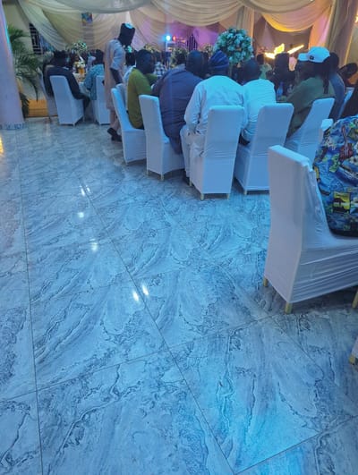 The image captures an indoor event, possibly a reception or gathering, with guests seated at tables covered by white cloths. The floor is tiled with a marble-like pattern in shades of blue and gray, reflecting ambient light. In the foreground, several chairs are draped with white slipcovers, and a portion of a person wearing a colorful Hawaiian-style shirt is visible on the right. In the middle ground, men are seated and standing, some wearing traditional African attire. The background features draped curtains and floral arrangements, suggesting a decorated venue. The overall lighting is dim with accents of blue and yellow. The image does not provide any specific landmarks or visual cues to definitively place the location in Barden Yana, Nigeria, although the attire of some individuals might suggest a Nigerian context.