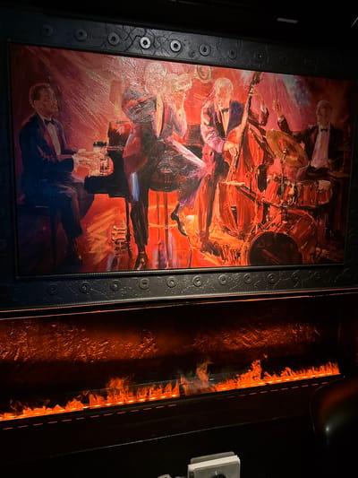 An oil painting depicting a jazz quartet playing in a dimly lit room hangs above a modern fireplace with artificial flames. The painting shows a pianist on the left, a bassist in the center, a drummer on the right, and another musician, possibly a saxophonist, partially obscured. The musicians are dressed in formal attire. The fireplace below the painting emits a warm, orange glow, creating a cozy atmosphere. The scene is indoors, likely in a restaurant, bar, or club. There is no information available to determine the specific location in Moscow, Russia.