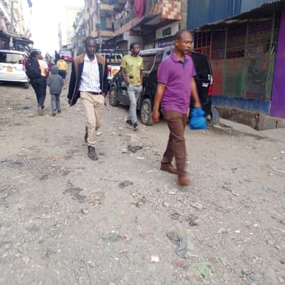 The scene depicts an unpaved, debris-strewn urban street in Nairobi, Kenya, illuminated by natural daylight. Six discernible individuals are present.

In the immediate foreground, a dark-complexioned adult male, wearing a light-colored striped shirt, a dark blazer, tan trousers, and dark boots, walks towards the camera. Adjacent to him on the right, another dark-complexioned adult male, dressed in a purple polo shirt, brown trousers, and brown shoes, walks away from the camera, carrying a small blue bag in his right hand. His movement is blurred, indicating motion.

In the midground, a third dark-complexioned adult male, wearing a light green t-shirt with a graphic design and light grey jeans, stands next to a parked black SUV. Behind this SUV, a white pickup truck is partially visible. Further left in the midground, an adult, possibly female, in dark clothing and carrying a backpack, walks away from the camera, preceded by a child in a grey jacket. A white minivan is parked on the far left of the street.

Multi-story buildings, consistent with residential or mixed-use commercial structures, line both sides of the thoroughfare. One building on the right features a partial sign reading "ST. CHRYSANTHEMUM" and exhibits clothes hanging on railings. Sections of the buildings are painted in various colors, including purple on the far right. The street surface is uneven, composed of dirt and gravel, and is littered with various refuse, including plastic bags, fabric scraps, and exhibits several dark, irregular stains.