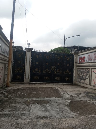 The image displays an exterior scene of a large property entrance in Bogor, Indonesia, under an overcast sky. A substantial, dark-colored metal gate, featuring ornate gold-colored scrollwork designs and pointed finials along its top, occupies the central view. The gate is supported by two light-colored pillars, each crowned with a decorative lantern-style light fixture. To the left, a light-colored perimeter wall shows black graffiti, including partially legible text "KISS XW" and "KB TO". To the right, a segmented perimeter wall is heavily marked with graffiti, notably "KBC XW" in red spray paint and multiple black scribbled tags; a single decorative light fixture is also mounted on its upper right section. A tall, dark utility pole stands vertically on the far left, partially wrapped in a lighter material near its base. Above the right side of the gate, a curved street lamp pole supports a horizontal luminaire. Thin utility lines are visible crossing the cloudy sky. The foreground comprises a grey concrete or asphalt driveway exhibiting extensive wear, stains, and patches of moisture. Green foliage from trees is visible behind the gate and walls. No people, vehicles, or specific actions are depicted.