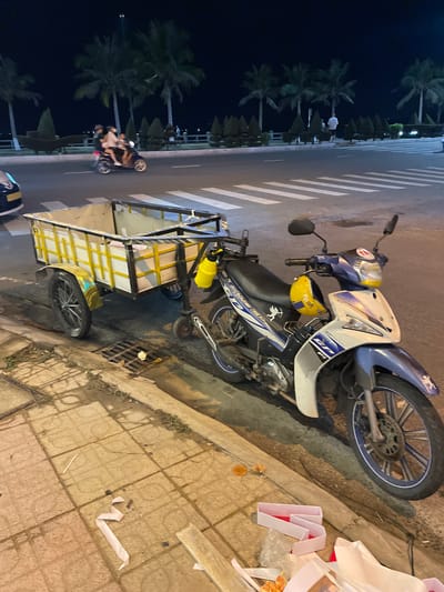The image shows a motorized tricycle parked on a paved sidewalk next to a street. The tricycle is hauling a large, yellow cargo box. The scene is set at night, with streetlights and the dark sky suggesting artificial lighting. Palm trees line the street in the background, indicating a tropical or subtropical location. A few people are visible on the street, either walking or riding other motorcycles. There are also some white and red packaging materials scattered on the sidewalk near the tricycle. The overall impression is of a street scene in an urban or semi-urban area in Vietnam at night.
There is some text visible on the motorcycle, but it is not legible in the image. The text on the yellow helmet reads "R p oy". The motorcycle itself has a blue and white color scheme with some graphic designs. The tricycle appears to be a common mode of transport for goods in the region. The lighting conditions suggest it is after sunset. The presence of palm trees and the style of the motorcycle are consistent with a location in Southeast Asia, specifically Nha Trang, Vietnam.