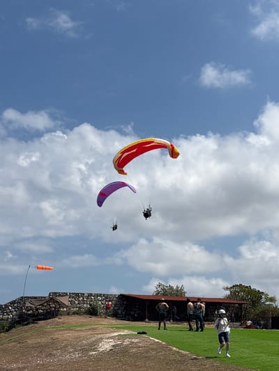 This image shows a paragliding launch site in Kuta Selatan, Indonesia. The sky is blue with scattered white clouds. Three paragliders are in the air. The largest paraglider, in the upper right, is red and yellow. Below and to the left of it, a purple paraglider is visible. In the distance, a smaller paraglider is also in the air. On the ground, a group of people are gathered near a building with a red roof. A windsock is visible on a pole to the left of the building. The ground is covered in dry grass with patches of green.