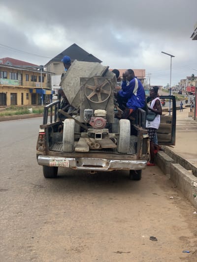 The scene shows a pickup truck parked on the side of a road in Akure, Nigeria (7.24, 5.197). The truck bed is loaded with a large, grey concrete mixer, two spare tires, wooden planks, and two metal pipes. Four men are sitting and standing on the truck bed, with one man in a blue shirt and one in a white shirt. The man in the blue shirt is sitting on the back of the truck, facing forward with his legs hanging off the side. The man in the white shirt is standing behind the concrete mixer, facing towards the camera. There is a light pole behind the truck with a street light attached to it. Other vehicles are visible in the background. The truck is parked on a dirt road in front of a concrete curb. The surrounding buildings are constructed of concrete and painted with a pale yellow color. The scene is bright and sunny with no signs of rain. The concrete mixer appears to be in working order.