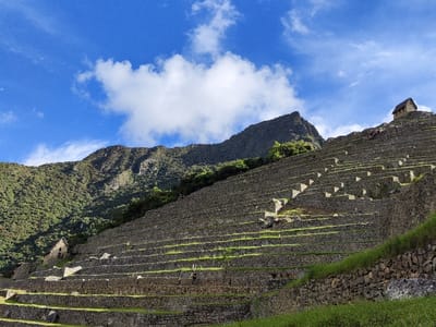 Machu Picchu's terraces rise dramatically against a blue sky dotted with clouds. The stone walls create steps towards the mountain peak, hosting a small structure. Lush green vegetation covers parts of the terraces and the slopes of the adjacent mountain. The detailed stonework and architectural design are characteristic of the ancient Inca civilization.
