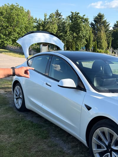 A white Tesla Model 3 electric car is parked on a grassy area next to a paved road. The trunk is open, revealing the interior. A person's left hand, wearing a black watch, is visible in the foreground, with their index finger pointing towards the car's rear passenger door. In the background, there are trees, a small wooden structure, and a fence. Beyond the fence, gravestones and crosses suggest the presence of a cemetery. The sky is clear and blue with a few white clouds. The image was taken in Luka, Ukraine.
