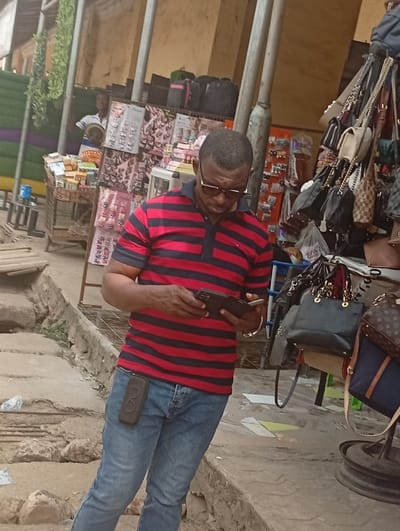 The image captures an outdoor market scene, likely during the daytime, with a focus on a man in the foreground. The overall atmosphere suggests a bustling commercial area, possibly a street market or a series of informal stalls.
In the center of the frame, a man with a dark complexion and short, closely cropped dark hair is prominent. He is wearing dark-framed sunglasses, a short-sleeved polo shirt with horizontal red and black stripes, and light blue denim jeans. His head is tilted downwards, and he is intensely focused on a dark-colored smartphone or similar device held open in both hands at waist height. A black, rectangular phone holster or case is clipped to the right side of his jeans. He also appears to be wearing a bracelet on his left wrist.
Behind the man, the background reveals multiple market stalls. To the left, a display rack holds numerous pairs of sunglasses, earrings, and other small accessories, suggesting a fashion accessory stall. Another individual, lighter-skinned and wearing a white shirt, can be partially seen peering from behind this stall in the upper left background. Further left, there's an area with green and purple colored panels or walls, adding a splash of color to the otherwise earthen tones.
To the right of the central figure, another stall is visible, heavily adorned with various handbags and purses of different sizes, colors, and styles, hanging from racks. One notable handbag features a distinctive monogram pattern, possibly indicating a designer brand replica. The ground in the foreground is uneven, appearing to be a mixture of dirt and rough concrete or paving stones, with some white plastic litter scattered about. Tall, unpainted metal poles support the overhead structures of the market stalls. The lighting is diffused, suggesting it's not direct midday sun, possibly late morning or early afternoon, with some areas appearing to be in partial shade. No clear, readable text or signage is visible in the image.