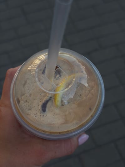 This is a top-down, close-up shot of a clear plastic cup filled with a frothy, light brown beverage, likely coffee or a similar drink. A white straw extends from the cup at an angle, disappearing into the drink. A thin, light yellow slice, possibly a lemon wedge, is visible floating on the surface of the drink, partially submerged. The drink has a creamy texture with small bubbles and dark specks. The lid of the cup is transparent, revealing the contents, and it has a small circular opening for the straw.
A person's hand is holding the cup from the right side of the frame. Their skin is visible, along with a fingernail painted a pale pink. The background is a textured, dark grey surface, appearing to be pavement made of interlocking blocks.
The lighting suggests it might be daytime, with ambient light reflecting off the surface of the drink and the plastic lid. The reflections on the lid show a pattern of square shapes, possibly from a building or an overhead structure, suggesting an outdoor setting, perhaps a street or a cafe patio. The overall mood is casual, possibly depicting someone enjoying a drink on the go. No visible text is present in the image. The context provided places the location as Brest, Belarus.