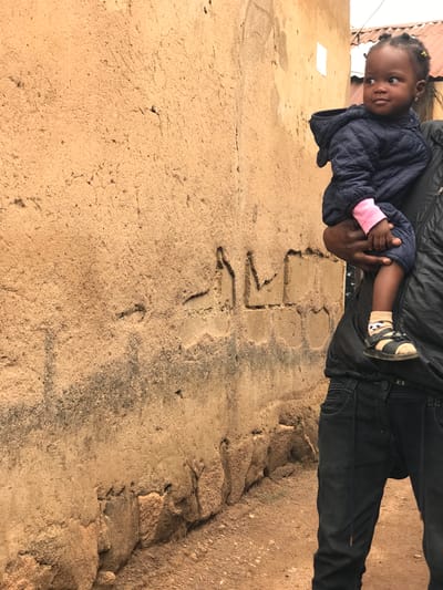 The image depicts a close-up view of a person carrying a young child in Kabong, Nigeria. The background is dominated by a textured, light brown wall, possibly made of mud or stucco, with some darker, weathered marks and visible structural imperfections. The ground in the foreground is sandy and reddish-brown, with small pebbles.
The child, who appears to be a toddler, is facing to the right and looking away from the camera. The child is wearing a dark blue, quilted hooded jacket with pink sleeves peeking out. The child's feet are clad in tan socks with a panda design and black sandals. The child is being held by a person whose body is partially visible on the right side of the frame. This person is wearing a black puffer vest over a dark shirt and dark blue jeans. Their hands are holding the child. The person's skin tone appears to be dark. The lighting suggests an overcast day.