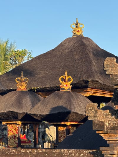 A low-angle, close-up outdoor shot features the traditional Balinese architecture of a temple in Ubud District, Indonesia. The roofs are thatched with dark, coarse material and topped with ornate, gold-colored crowns. One crown is prominently placed on the highest roof, a conical structure dominating the center of the frame. Two smaller, similar crowns adorn the peaks of adjacent, lower thatched roofs in the foreground. These roofs are supported by intricate wooden structures with gold and red carvings, visible beneath the overhangs. In the lower portion of the image, stone steps lead up to a raised platform where ornate golden sculptures and red pedestals are visible. The stone detailing on the right side of the frame suggests part of a temple wall or entrance. The sky above is a clear, bright blue, indicating a sunny day. The lighting suggests it is either early morning or late afternoon, casting long shadows and highlighting the golden decorative elements. There are no people visible in the image.
