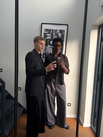 Two young adult males are visible in a full-length mirror selfie, positioned in a modern indoor setting identified as being in the city of London, United Kingdom, evidenced by the presence of a British Standard BS 1363 electrical outlet on the right wall.
The subject on the left, with light skin and short, curly blonde hair, wears a black suit jacket over a black collared shirt, paired with black wide-leg trousers and black shoes. A silver stud earring is visible on his left ear, and a silver chain bracelet adorns his left wrist. He holds a dark blue smartphone in his right hand, angled towards the mirror.
The subject on the right, with dark skin and dark, curly hair, wears yellow-tinted aviator-style sunglasses. He is dressed in an unbuttoned brown long-sleeved collared shirt over a dark undershirt, grey wide-leg trousers, and black shoes. Multiple silver necklaces are visible around his neck, and silver rings adorn his fingers. He holds a light-colored smartphone in his left hand, also angled towards the mirror.
The background consists of a plain white wall. To the left of the subjects, a black metal industrial-style staircase with diamond plate treads descends. Above and behind the subjects, a black-framed artwork with black and white abstract or text-based design is mounted on the wall. The floor is covered with a textured, medium brown carpet. The mirror itself is framed in black. To the far right, a vertical black frame suggests a window or glass door.