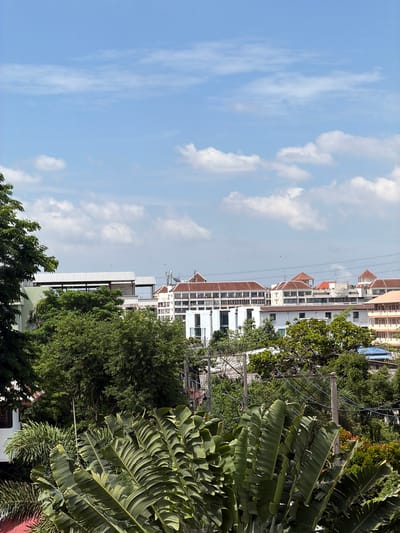 The image captures a daytime view of a city skyline in Bangkok, Thailand, characterized by a blend of lush greenery and urban architecture under a partly cloudy sky. In the foreground, large, tropical banana leaves dominate the lower portion of the frame, their broad, ribbed surfaces angled upwards and towards the viewer, creating a naturalistic curtain. Behind these leaves, a dense canopy of various green trees forms a middle ground, interspersed with the tops of several buildings.
The buildings are a mix of styles, suggesting a developed urban area. Some structures feature white facades and modern architectural lines, while others display traditional terracotta-tiled roofs with peaked designs, typical of many buildings in Southeast Asia. One notable building has a distinctive white, angled roof structure. Power lines and utility poles are visible throughout the middle ground, indicating the presence of essential infrastructure.
Further in the distance, the city expands with more buildings of varying heights and designs. The sky above is a bright blue, scattered with fluffy white cumulus clouds, hinting at fair weather. There are no discernible people or active human subjects in the frame; the scene is a static depiction of the urban and natural landscape. The overall impression is one of a bustling city where nature and development coexist, viewed from a slightly elevated perspective.