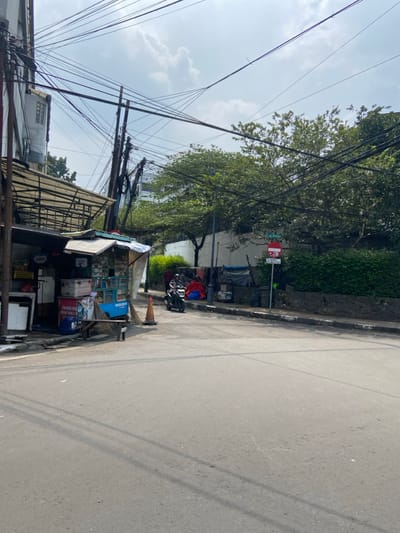 The scene is a street corner in Bandung, Indonesia, located at -6.923 latitude and 107.61 longitude. The street is paved and appears to be relatively empty, with a single motorcycle parked at the corner. There are a few people walking in the distance and a small shop on the corner. The street is lined with trees, some of which have branches reaching over the street. There are many power lines overhead. The sky is partly cloudy.