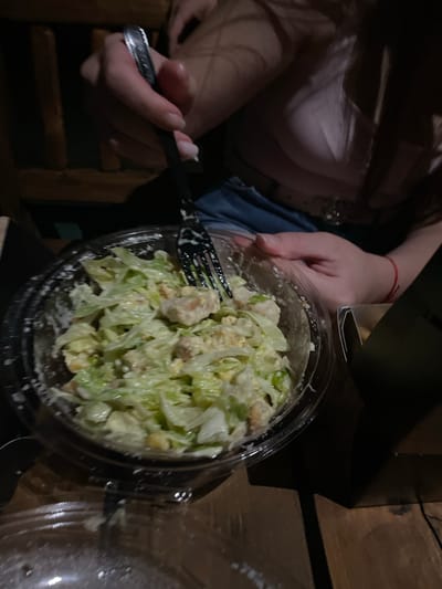 A close-up shot shows a person's hands holding a fork and a plastic container of salad. The salad appears to be a Caesar salad with lettuce, croutons, and chicken. The person is wearing a pink shirt and blue jeans. The background is dark and blurry, but it appears to be an indoor setting with wooden furniture. The lighting is dim. The image does not provide any specific location information.