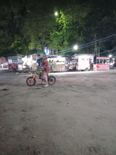 The image depicts an outdoor night scene in Nganjuk, Indonesia, likely within a public park or market area, illuminated by artificial lights. The foreground consists of an uneven, unpaved dirt or gravel surface.
Centrally positioned in the mid-ground, a person, appearing to be an adult, stands next to a small, customized motorbike. The individual wears a reddish-brown long-sleeved garment and light-colored sandals. The motorbike features yellow front forks, red wheel hubs, and knobby tires, with a visible green and yellow color scheme on its frame or tank. The person is oriented towards the left, appearing to be standing with or pushing the bike.
Behind the person and extending to the right, multiple food stalls and carts are visible. From left to right:
* A rectangular cart with a light green canopy and a sign partially visible, suggesting "ES SARI TEHUM" (Iced Tea Concentrate).
* Further right, a brightly lit white cart or small vehicle.
* To the far right, a stall with a prominent red banner advertising "SOSIS BAKAR JUMBO" (Grilled Jumbo Sausage), "TELOR GULUNG" (Rolled Egg), and "KENTANG GORENG" (French Fries).
Additional indistinct stalls and figures are present behind these main structures.
The background features dense foliage from trees, some brightly lit by overhead streetlights, and power lines are visible spanning across the upper frame. The overall lighting is a mix of bright localized illumination from the stalls and streetlights against a dark night sky.