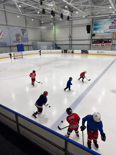 An indoor ice hockey rink in Votkinsk, Russia, is shown from a high-angle perspective. Six young hockey players in full gear are on the ice. Four players are actively engaged in skating or practicing, while two are further away, possibly waiting their turn. The rink has standard markings and a goal net is visible in the background. Large banners with text in Russian are displayed along the arena walls. The ceiling features exposed metal structures and lighting fixtures. The overall scene depicts a youth hockey training session.