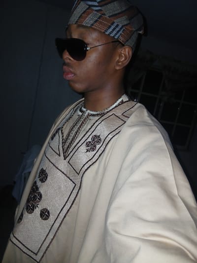 A medium shot shows a young Black person from the chest up, posed in profile to the left. They wear sunglasses, a traditional dashiki with intricate brown and gold embroidery on the chest and sleeves, and a patterned cap. The background is dark, with a window frame visible in the upper right corner. The image is likely a casual portrait taken indoors, possibly in a residential setting in Lagos, Nigeria, given the attire.