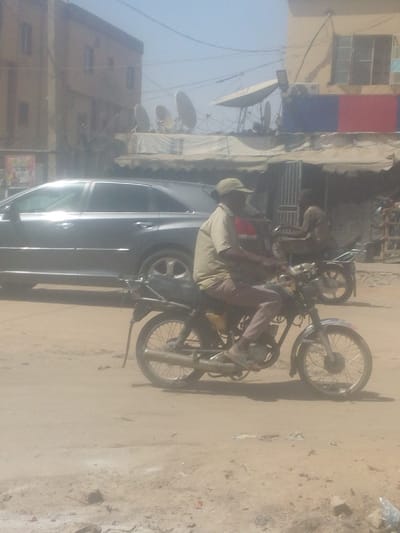 This image captures a street scene in Sabon Gari West, Nigeria. The foreground and midground consist of a dusty, unpaved road.

In the midground, a dark-skinned male is seated on a black motorcycle, facing right. He wears a tan short-sleeved shirt, tan pants, and a tan baseball cap. His hands are on the handlebars, and his feet are on the footrests. The motorcycle has spoked wheels, a chrome front fender, and an exposed exhaust pipe. A yellow container is visible near the engine. Behind him, a dark-colored sedan, possibly a Toyota Venza, is visible, oriented in the same direction. Further right and slightly behind, a second, partially obscured dark-skinned individual, appearing younger, is seated on another motorcycle near a metal gate.

The background features several buildings under a hazy sky. To the left, a multi-story, light-brown building with multiple rectangular windows is visible. To the right, a building displays a lighter upper section and a lower facade with horizontal red and blue panels. An awning extends from this building over the street level. On the rooftop, or an adjacent structure, at least five large satellite dishes are visible, along with exposed wiring and what appears to be an air conditioning unit. Overhead utility wires traverse the upper portion of the frame. The ground shows scattered debris, including plastic remnants.