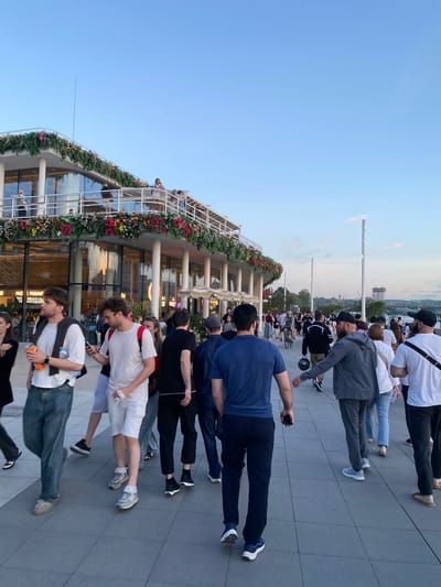 The image shows a lively scene on a pedestrian promenade in Moscow, Russia. In the background, a modern building is adorned with vibrant floral decorations, suggesting a festive occasion or a well-maintained public space. People are seen walking in various directions along the wide, paved promenade. The sky is clear blue, indicating a pleasant day. Several individuals are captured in motion, with some holding drinks or phones, while others are engaged in conversation or simply enjoying the stroll. The overall atmosphere appears to be one of leisure and social gathering.
