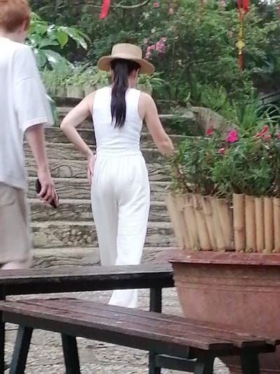 The image shows two people outdoors. On the left, a person wearing a white t-shirt and beige shorts is partially visible from the side. They are holding a dark-colored mobile phone in their right hand.
The main subject, to the right, is a person from behind, wearing a white sleeveless top and white wide-leg pants. They have a straw hat on their head and dark hair pulled back in a ponytail. Their left hand is on their hip, and their right arm is extended slightly as if reaching for something or posing.
They are standing on a stone staircase in a lush, green environment. To the right of the stairs is a large planter filled with green plants and some bright pink flowers. The planter itself is made of bamboo and sits in a larger reddish-brown pot. In the foreground, a dark wooden bench is visible. Red and gold decorative ribbons hang in the background amongst the foliage, suggesting a festive or cultural context.
The scene appears to be a garden or park, likely in the daytime given the natural lighting. The overall atmosphere seems relaxed and pleasant. There is no visible text in the image.