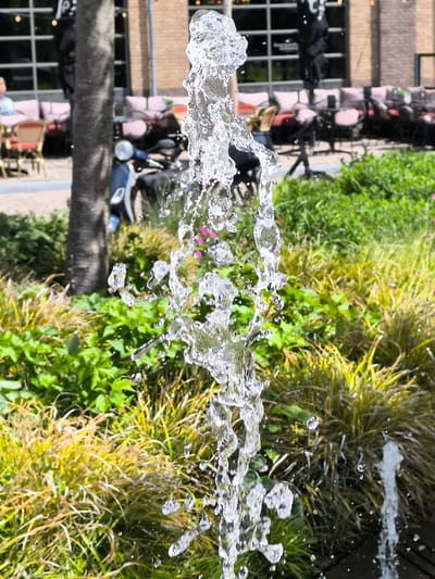 A close-up, vertical shot captures a vibrant stream of water from a fountain, frozen mid-air, with countless individual droplets glistening in the sunlight. The clear water forms an irregular, dynamic column, its texture and motion emphasized by the shallow depth of field.
In the immediate foreground and mid-ground, a lush array of plants and bushes provides a natural setting. These include rich green foliage and patches of drier, yellowish-green grasses, creating a textural contrast around the water feature. To the bottom right, another smaller jet of water is visible, indicating part of a larger fountain installation.
The background is softly blurred, revealing an urban setting. A dark tree trunk stands prominently on the left. Behind it, and extending across the mid-ground, is an outdoor cafe or restaurant. Faintly visible are tables with chairs, some featuring red or pink cushions, suggesting a comfortable dining area. A blue scooter or moped is parked near the tree, partially obscured. The facade of a building with large windows can be seen further back, and indistinct figures, likely patrons, are present at the cafe tables.
The scene is brightly lit, indicating a sunny daytime. The overall atmosphere appears pleasant and relaxed, typical of a European town square or pedestrian area. No distinct text is legible within the image. This scene is set in Alkmaar, Netherlands.