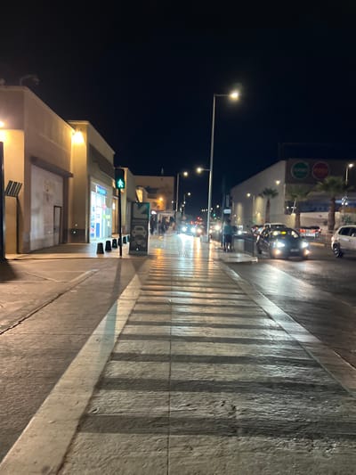 The image depicts a crosswalk at night in the city of Arica, Chile. Streetlights illuminate the scene, casting long shadows from the pedestrian crossing onto the wet pavement. Buildings line the street, some with glowing windows and awnings. Several cars are visible, with headlights on, navigating the road. A traffic light shows a green pedestrian signal. A signpost with an arrow indicating "ACCESO" (access) for cars and motorcycles is also present. The overall atmosphere is that of a well-lit urban street after dark.