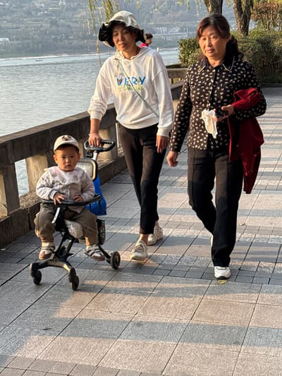 A group of three individuals, consisting of two adult females and one toddler, are walking on a paved riverside promenade in Jiangjin, China. The scene is illuminated by strong, warm sunlight from the left, casting long shadows to the right across the pathway.

The toddler, an East Asian child estimated to be 1-2 years old, is seated in a black-framed, multi-wheeled stroller. The child wears a light-colored padded jacket with a patterned collar, khaki pants, brown and white sneakers, and a light-colored baseball cap with an animal face design. The child faces forward with a neutral expression.

An adult East Asian female, appearing middle-aged, pushes the stroller with her right hand. She wears a white long-sleeved hooded sweatshirt with a colorful graphic print, black pants, white sneakers, and a light-colored bucket hat with a dark underside. A light-colored strap crosses her torso.

A second adult East Asian female, appearing older middle-aged or elderly, walks to the right and slightly ahead of the first woman. She wears a black long-sleeved collared shirt with a white collar and small light patterns, black pants, and white sneakers. She carries a folded red jacket over her left forearm and holds a white cloth or tissue in her right hand.

The pathway is composed of light gray rectangular paving tiles. A dark-colored metal railing separates the path from a wide body of water, likely a river, where a small boat is visible in the distance. The opposite bank features distant hills or mountains, scattered buildings, and a visible bridge structure. Sparse green foliage is present on the right side of the pathway.