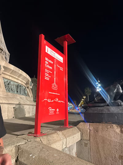 The image captures a night scene in Barcelona, Spain, focusing on a prominent red sign for "Mirador de Colom" (Columbus Viewpoint) and "Official Tourist Information." The sign details services like "Official Tickets & Tours," "Unique experiences," and "Personal attention," and displays "Panoramic Views." Below this, a website address "tickets.visitbarcelona.com" is visible, along with the Barcelona Turisme logo. To the left of the sign, a portion of a large monument, possibly the Columbus Monument itself, is visible, featuring detailed relief sculptures. The background shows a street with lit lampposts and some distant traffic lights, suggesting an urban environment. A dark, shadowy statue or sculpture is visible in the lower right of the frame, partially illuminated by what appear to be light beams. The ground consists of rough stone blocks.