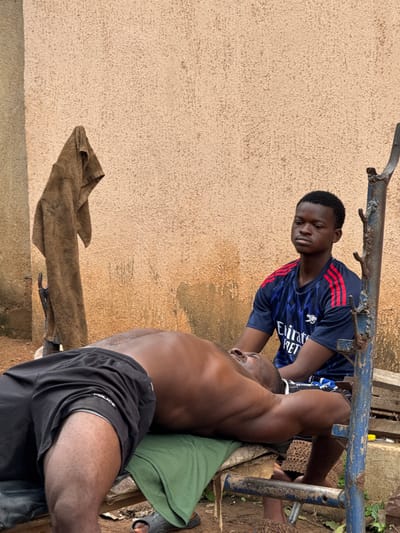 In Akwanga, Nigeria, two young men are engaged in a weightlifting activity outdoors. The man on the bench is lying on his back, lifting a weight which is not visible in the frame. The other man, seated, appears to be assisting or spotting him. The setting is rustic, with a textured, light-colored wall forming the backdrop. A brown towel hangs on a pole to the left of the frame, and a blue metal bench is utilized for the exercise. The ground is dirt and scattered with debris. The focus is on the physical exertion and the interaction between the two individuals.