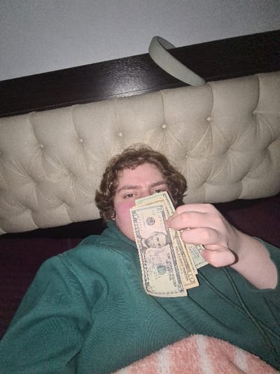 A person lies on a bed or couch, holding a stack of US dollar bills partially obscuring their face. The person wears a green sweatshirt. The couch has beige, tufted upholstery, and is positioned against a dark wood frame and a light gray wall. The money appears to be a mix of five and ten dollar bills.