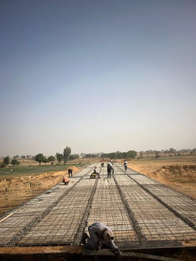 The image depicts a construction site, likely for a large concrete structure such as a road, canal, or bridge deck, extending into the distance under a hazy sky. The perspective is from an elevated position, looking down and along the length of the structure.
In the foreground and extending along the length of the structure, there is a grid of metal rebar (reinforcing steel bars) laid out, ready for concrete pouring. This rebar forms several parallel lanes or sections. On the far right, there's a visible concrete edge or formwork.
Several workers are present on the site, engaged in various activities related to the rebar installation and preparation.
* In the immediate foreground, on the far right edge, a worker wearing light-colored traditional attire and a hat is crouching, appearing to work on the formwork or the edge of the rebar grid.
* Further along the structure, towards the middle ground, multiple workers are standing or moving between the rebar grids. Some appear to be inspecting the rebar, others might be tying it. One worker closer to the center-right is seen operating what looks like a small piece of machinery or equipment on the rebar grid.
* In the background, more workers are visible along the length of the structure, suggesting a widespread construction effort.
The surrounding environment consists of dry, sandy-colored earth, indicating a possibly arid or semi-arid region. There are scattered green trees and some patches of cultivated land (possibly fields) visible on either side of the construction path, particularly in the middle and far distance. The sky is very light blue to almost white, suggesting a bright, possibly hot, and hazy day. There are no visible clouds.
No discernible text is visible in the image. The overall impression is one of active, large-scale infrastructure development in a rural or undeveloped setting.