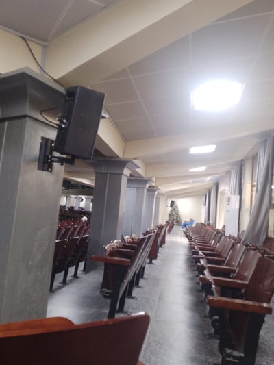 The image shows a wide view of the interior of a hall, likely an auditorium or conference room, in Kuson, Nigeria. The hall features rows of dark wooden chairs with dark metal legs, arranged in long aisles. The chairs appear to be fixed seating. On the left side of the image, a large, dark gray pillar is visible, mounted with a black speaker system. The ceiling is a suspended grid system with recessed lighting panels, one of which is brightly illuminated. Drapes hang along the right wall, and an air conditioning unit is visible near the right wall. Further down the hall, the seating continues, and in the distance, there is a decorated Christmas tree. A few people are visible scattered throughout the seating area. The overall environment suggests a formal gathering or event space.