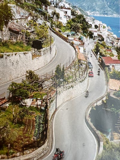 This media file depicts a scenic, winding road carved into a hillside with lush greenery and buildings clinging to the slopes. The road is paved with cobblestones in some sections and asphalt in others. Several vehicles, including cars and motorcycles, are visible on the road, suggesting a race or a tour.
In the foreground, a motorcycle with a rider is in motion. Further up the road, a red car and several other vehicles are also present. The overall scene is vibrant and bathed in natural light, suggesting it is daytime. The architecture of the buildings and the vegetation hint at a Mediterranean or similar climate. The presence of the winding road and the buildings cascading down the hillside suggest a location with dramatic topography, possibly a coastal town or a mountainous region. There is no visible text in the image.